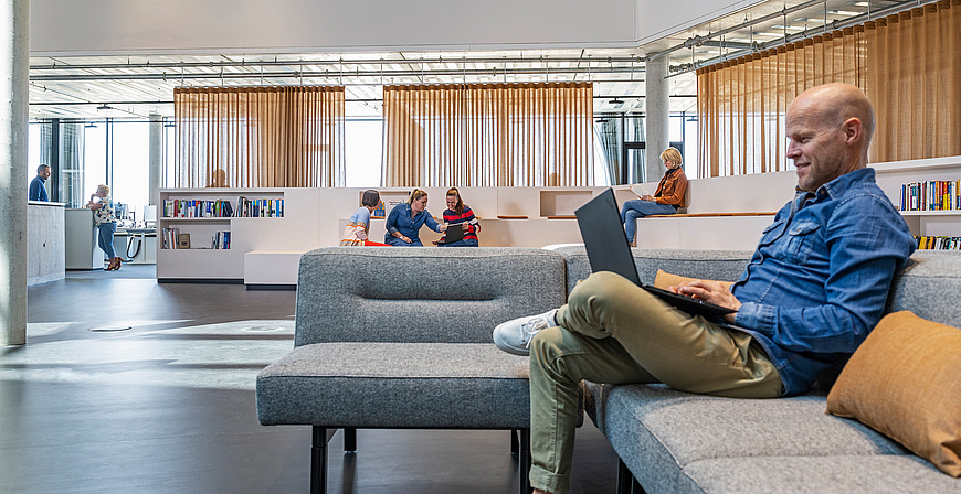 Ein Mitarbeiter arbeitet auf einer grauen Couch im offenen Bürobereich mit weiteren Kollegen im Hintergrund