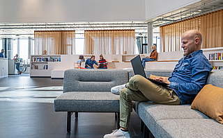 Ein Mitarbeiter arbeitet auf einer grauen Couch im offenen Bürobereich mit weiteren Kollegen im Hintergrund