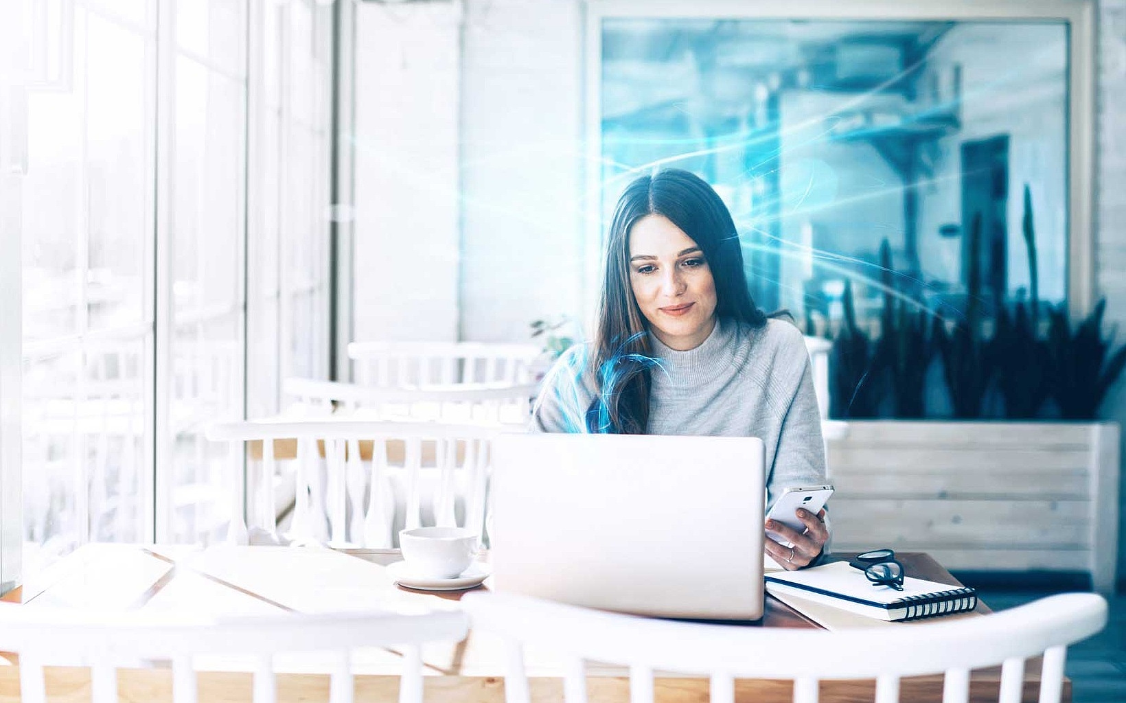 Frau sitzt in einem Café und arbeitet an ihrem Laptop, während sie ihr Smartphone in der Hand hält