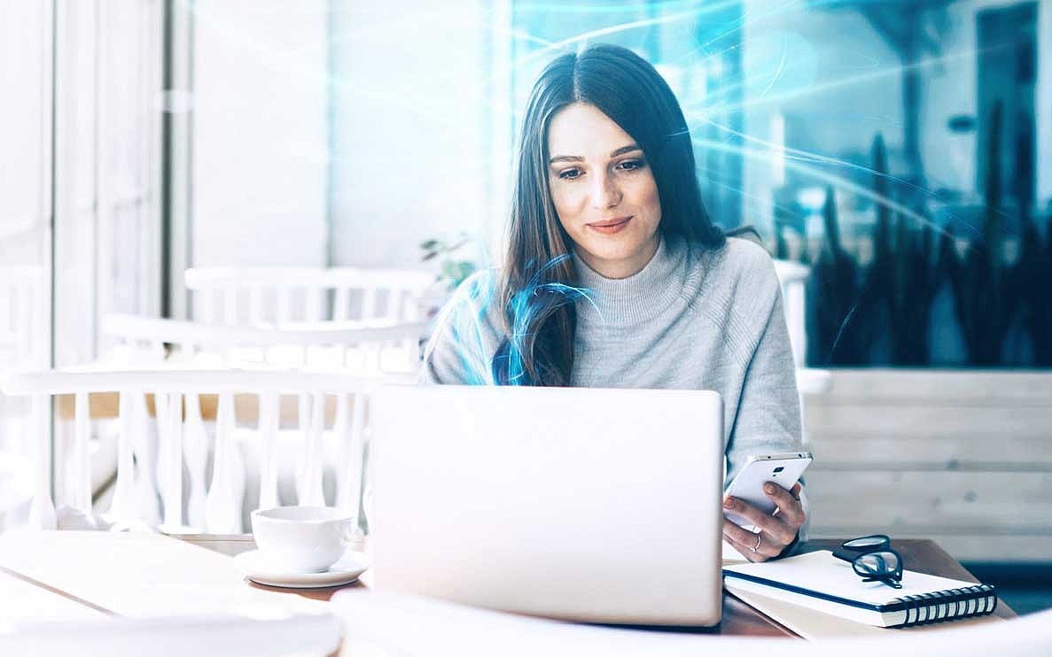 Frau sitzt in einem Café und arbeitet an ihrem Laptop, während sie ihr Smartphone in der Hand hält