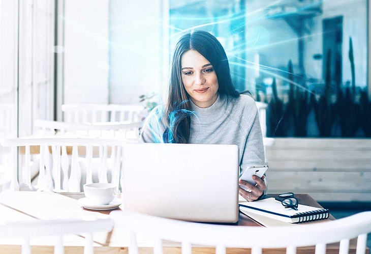 Frau sitzt in einem Café und arbeitet an ihrem Laptop, während sie ihr Smartphone in der Hand hält