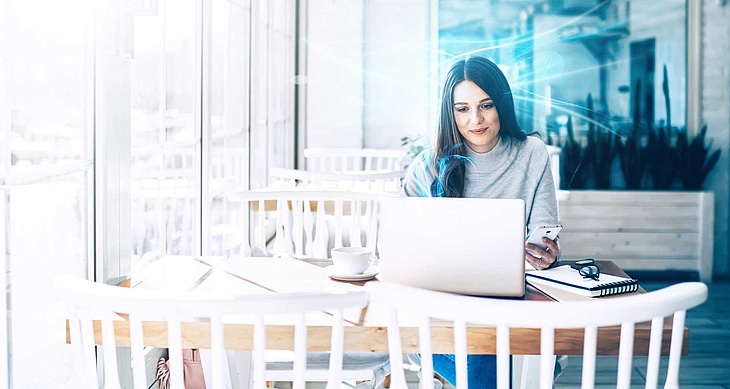 Frau sitzt in einem Café und arbeitet an ihrem Laptop, während sie ihr Smartphone in der Hand hält