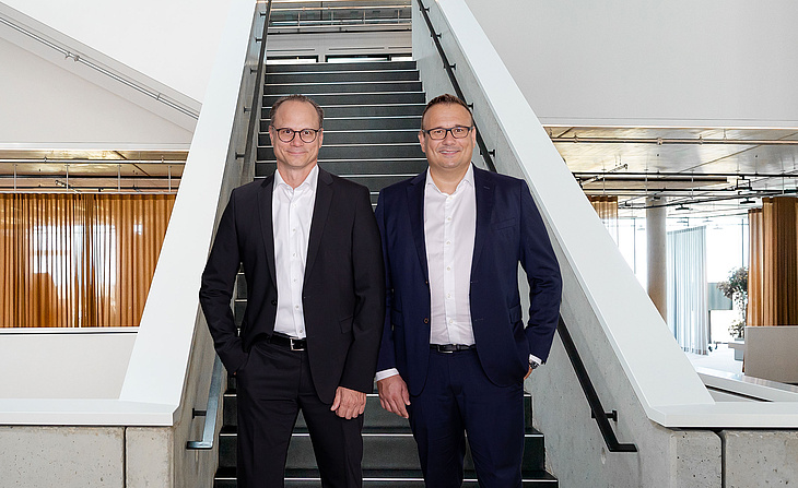 Die Geschäftsführer Michael Huber und Lars Rippert von baramundi stehen vor einer Treppe in einem modernen Bürogebäude